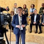 Un hombre habla frente a una cámara con un grupo de personas detrás en un espacio moderno.