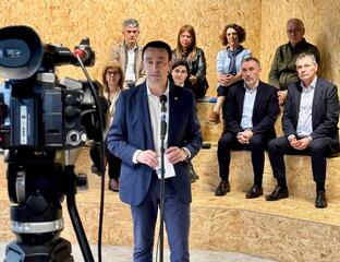 Un hombre habla frente a una cámara con un grupo de personas detrás en un espacio moderno.