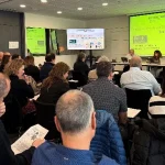 Reunión con participantes en un salón moderno, pantallas visibles.