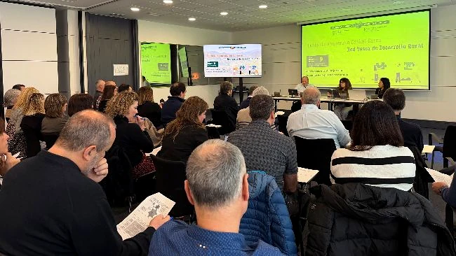 Reunión con participantes en un salón moderno, pantallas visibles.