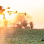 Hombre con sombrero observa un dron sobre un campo al atardecer.