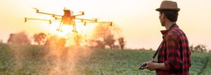 Hombre con sombrero observa un dron sobre un campo al atardecer.