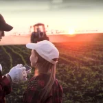 Dos personas en un campo con tractor al fondo durante el atardecer.