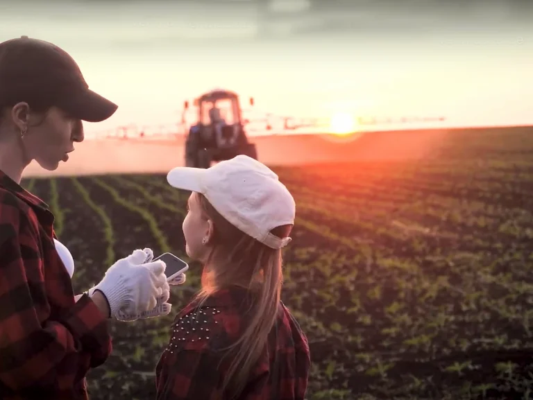 Dos personas en un campo con tractor al fondo durante el atardecer.